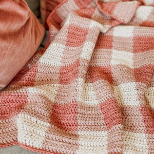 May include: A soft, crocheted throw blanket in a pink and white gingham pattern. The blanket is made with a chunky yarn and has a relaxed, cozy feel.