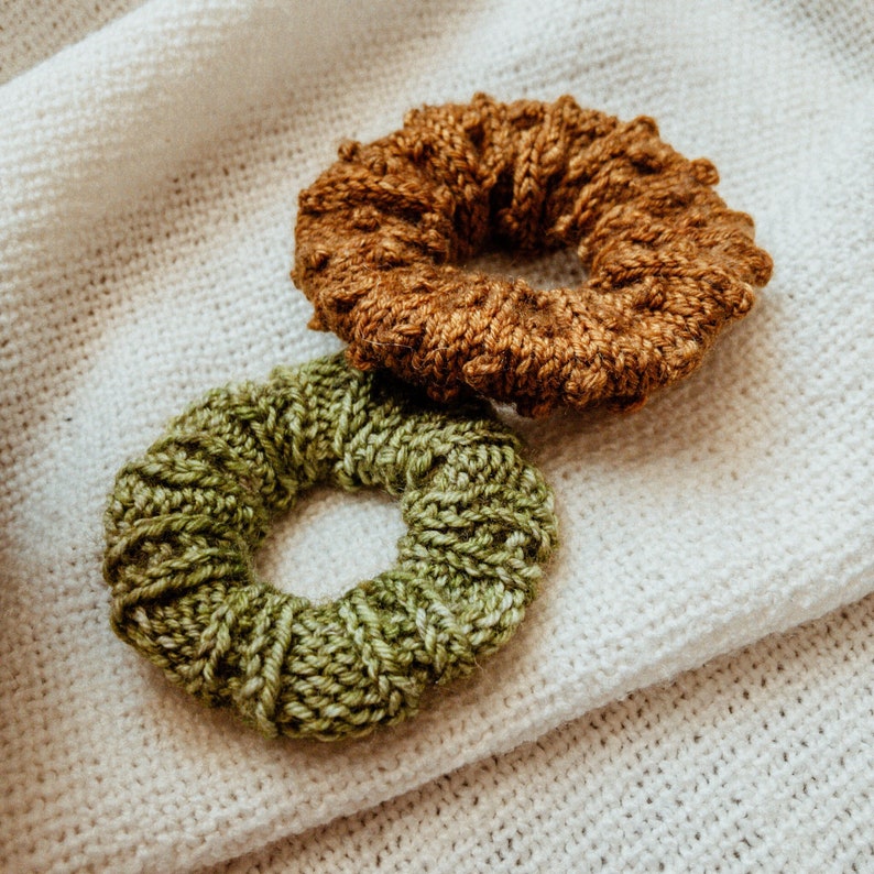 K&ouml;nnte beinhalten: Zwei geh&auml;kelte Haargummis, eines braun und eines gr&uuml;n, auf einem wei&szlig;en gestrickten Stoffhintergrund.