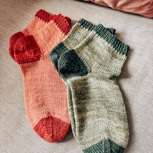 K&ouml;nnte beinhalten: Zwei Paar gestrickte Socken, eine in einem rosa-roten Farbschema und die andere in einem gr&uuml;n-grauen Farbschema.