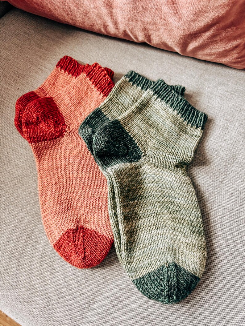 K&ouml;nnte beinhalten: Zwei Paar gestrickte Socken, eine in einem rosa-roten Farbschema und die andere in einem gr&uuml;n-grauen Farbschema.