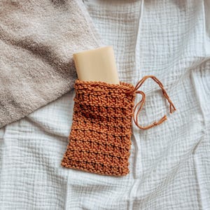 May include: A brown knitted soap sack with a white bar of soap inside. The sack has a drawstring closure and is lying on a white textured fabric.