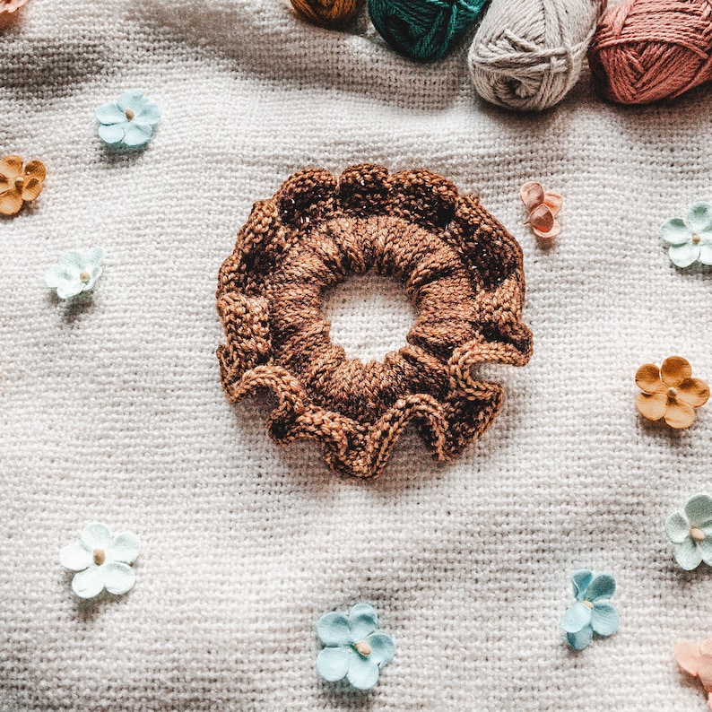 May include: A brown knitted hair scrunchie with a ruffled edge. The scrunchie is lying on a white knitted fabric background.