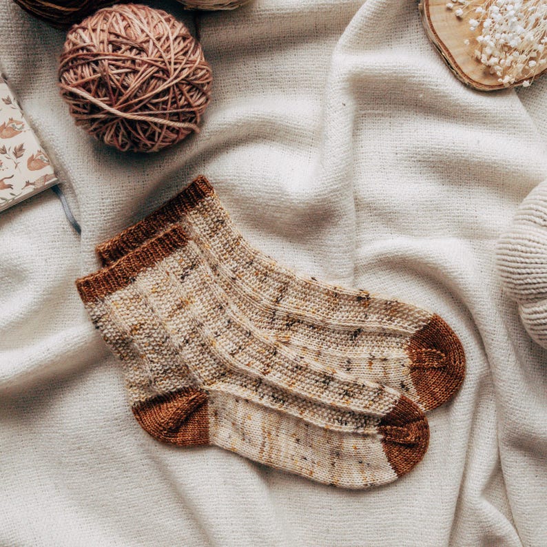 May include: A pair of knitted socks in a beige and brown color scheme with a speckled pattern. The socks have a brown cuff and are folded over.