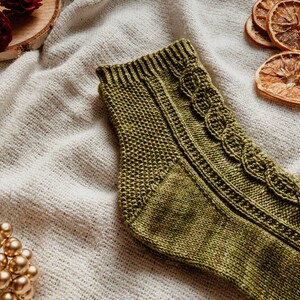 May include: A green knitted sock with a cable knit pattern. The sock is lying on a white fabric background.