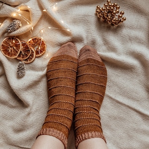 Puede incluir: Un par de calcetines de punto a rayas marrones y beige sobre un fondo de tela blanca con luces de cadena y rodajas de naranja secas.