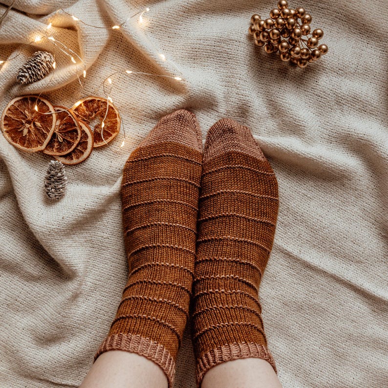Puede incluir: Un par de calcetines de punto a rayas marrones y beige sobre un fondo de tela blanca con luces de cadena y rodajas de naranja secas.