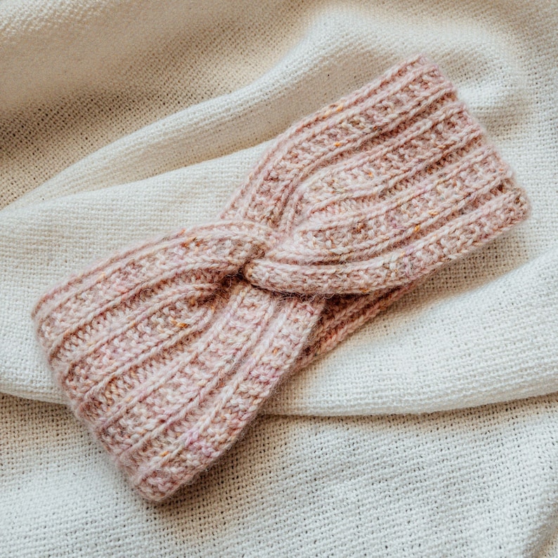 May include: A pink knitted headband with a twisted knot design. The headband is made of a soft, textured yarn.