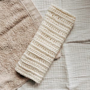 May include: A white crocheted washcloth with a textured pattern. The washcloth is folded and placed on top of a beige towel.