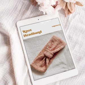 May include: A digital tablet displaying a knitting pattern for a knotted headband. The pattern is titled "Knot Headband" and the website is "wjjleberry". The headband is a light pink color.