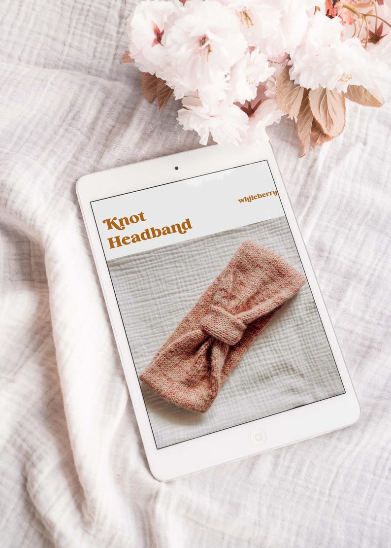 May include: A digital tablet displaying a knitting pattern for a knotted headband. The pattern is titled "Knot Headband" and the website is "wjjleberry". The headband is a light pink color.