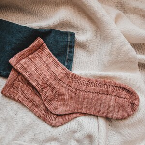 May include: A pair of pink knitted socks with a ribbed texture. The socks are lying on a white fabric background.