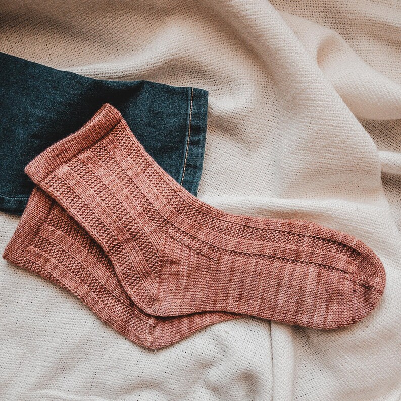 May include: A pair of pink knitted socks with a ribbed texture. The socks are lying on a white fabric background.