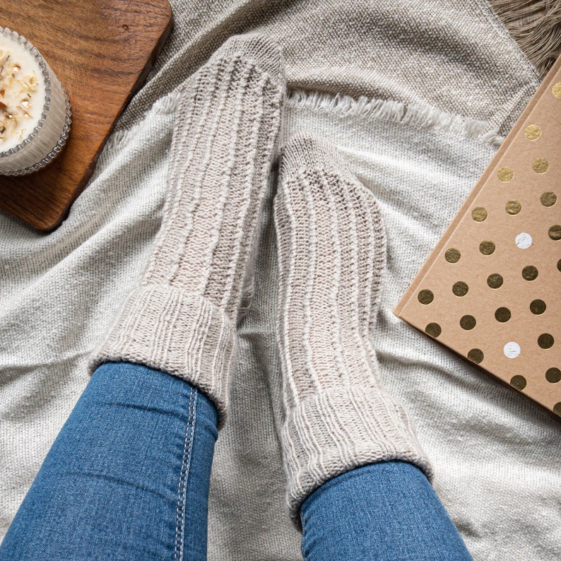 May include: A pair of light beige ribbed knit socks with folded cuffs. The socks are worn with blue jeans. A candle in a glass jar and a gift box with gold polka dots are also visible.