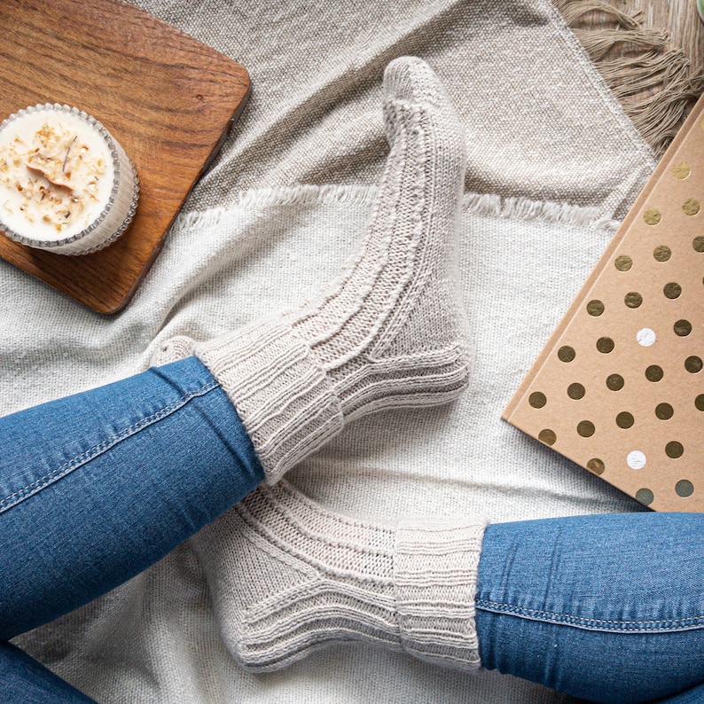 May include: A pair of light gray knitted socks with ribbed and patterned sections. The socks are worn with blue jeans. A wooden tray with a candle and a gift box with gold polka dots are also visible.