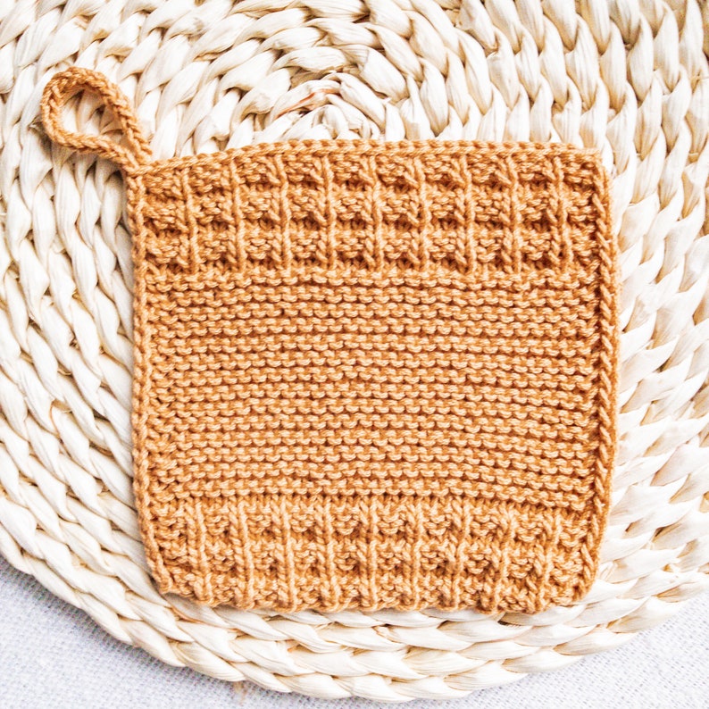 May include: A knitted dishcloth in a warm brown color with a textured pattern. The dishcloth has a loop handle and is made of cotton yarn.
