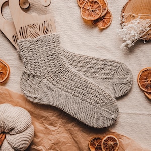 May include: A pair of knitted beige socks with a ribbed texture. The socks are laid out on a beige fabric background with dried orange slices and a small white pumpkin.