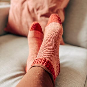 Peut inclure: Une paire de chaussettes en tricot dans des tons de rose et de rouge, avec les chaussettes remontées jusqu'aux chevilles. Les chaussettes reposent sur un canapé gris clair.
