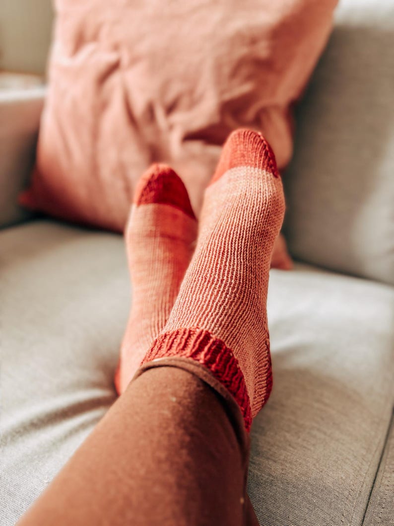 K&ouml;nnte beinhalten: Ein Paar gestrickte Socken in Rosa- und Rott&ouml;nen, wobei die Socken bis zu den Kn&ouml;cheln hochgezogen sind. Die Socken liegen auf einem hellgrauen Sofa.