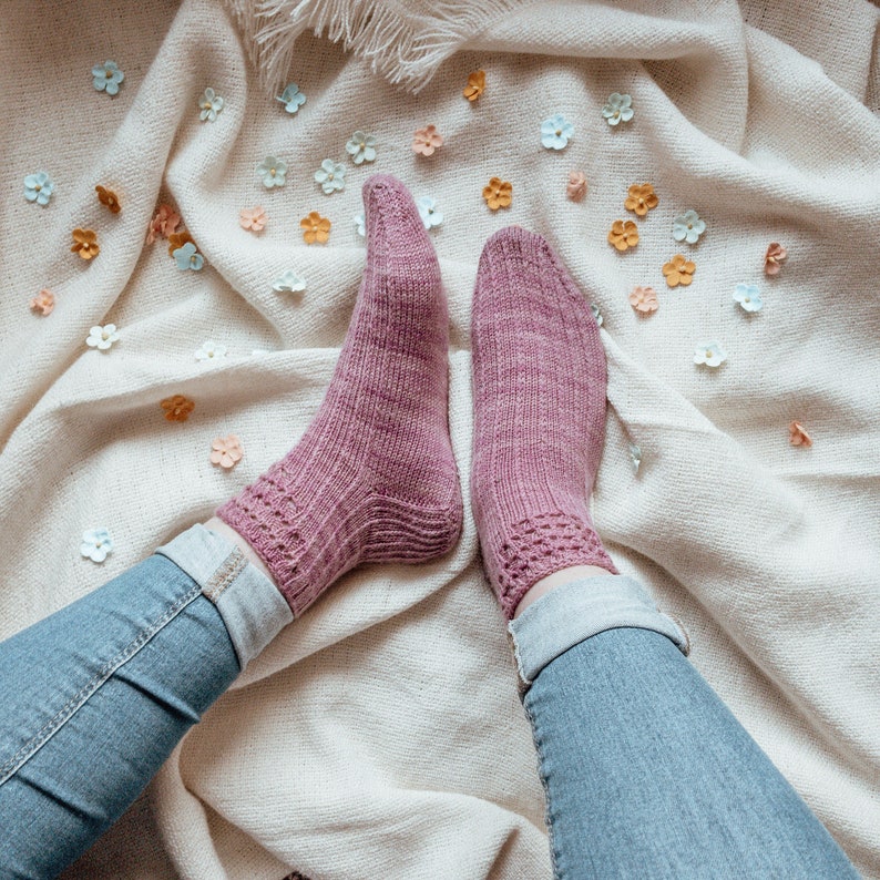 May include: A pair of knitted socks in a dusty rose color. The socks have a ribbed texture on the foot and a decorative openwork pattern at the cuff. The socks are worn with blue jeans, resting on a cream-colored blanket scattered with small, colorful paper flowers.
