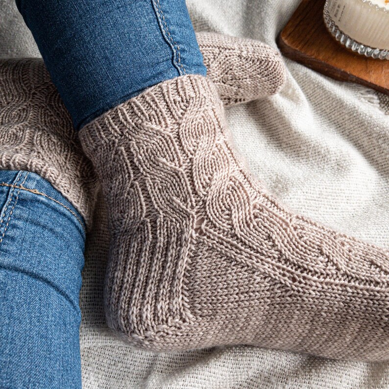 May include: Close-up of beige cable-knit socks. The socks are worn with blue jeans. The socks have a detailed cable pattern on the front and top of the foot. The socks appear to be handmade and are a neutral color.