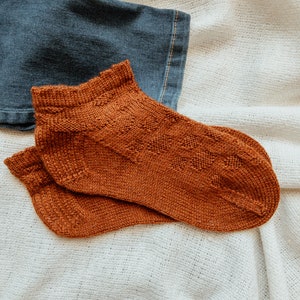 May include: A pair of rust-colored knitted ankle socks with a diamond pattern. The socks are made of a soft, textured yarn and are displayed on a white, textured surface next to a piece of blue denim.