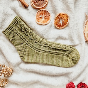 May include: A green knitted sock with a cable pattern on the top and a textured knit on the bottom. The sock is lying on a white fabric background.