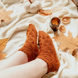 May include: A pair of rust-colored knitted socks with a textured pattern, resting on a cream-colored blanket. The scene includes a small white pumpkin, dried orange slices, a lit candle in a glass jar, and autumn leaves, creating a cozy fall aesthetic.