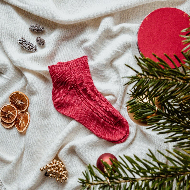 May include: A red knitted sock with a cable knit pattern on a white background with pine branches, dried orange slices, and gold ornaments.