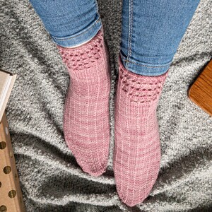 May include: A pair of pink knitted socks with a patterned cuff, worn with blue jeans. The socks are a soft, muted pink with a textured appearance. The cuff features a decorative openwork design.