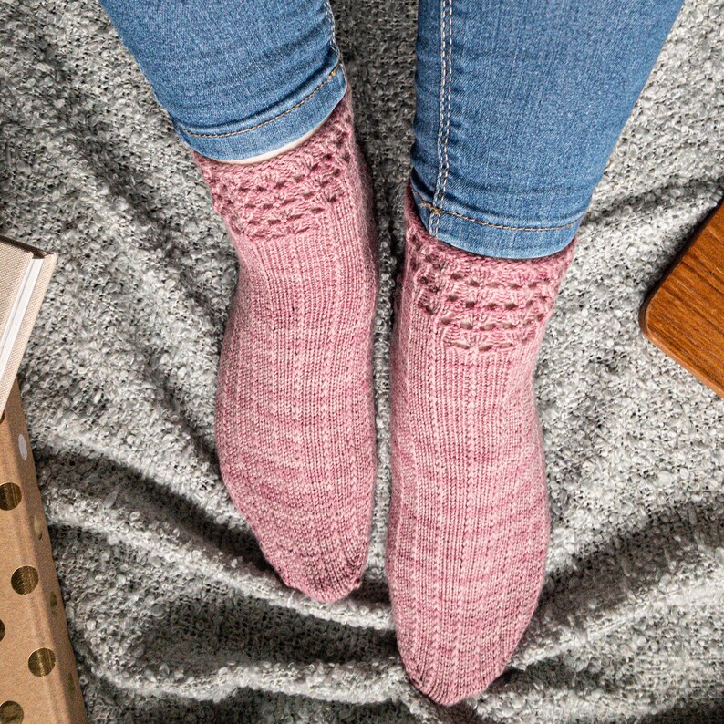 May include: A pair of pink knitted socks with a patterned cuff, worn with blue jeans. The socks are a soft, muted pink with a textured appearance. The cuff features a decorative openwork design.