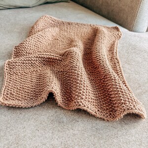 Op de afbeelding: Een rechthoekige, lichtbruine gebreide doek met een consistent patroon in ribbelsteek. De stof heeft een getextureerd, bobbelig oppervlak dat wordt gecre&euml;erd door de in elkaar grijpende lussen van garen, met afgewerkte randen.