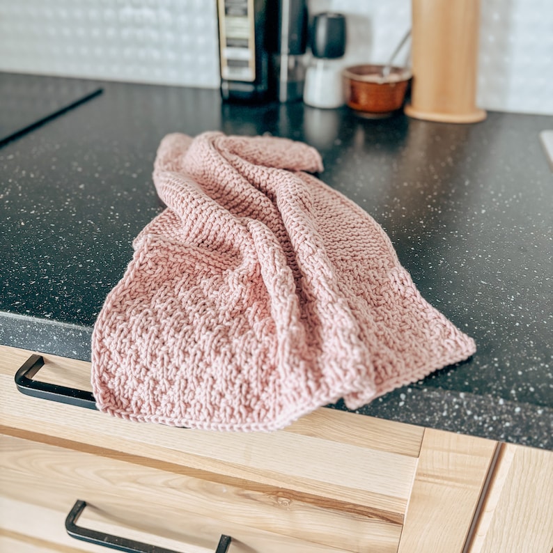 May include: A pink knitted dishcloth or tea towel on a black countertop. The cloth is folded and has a soft, textured surface.