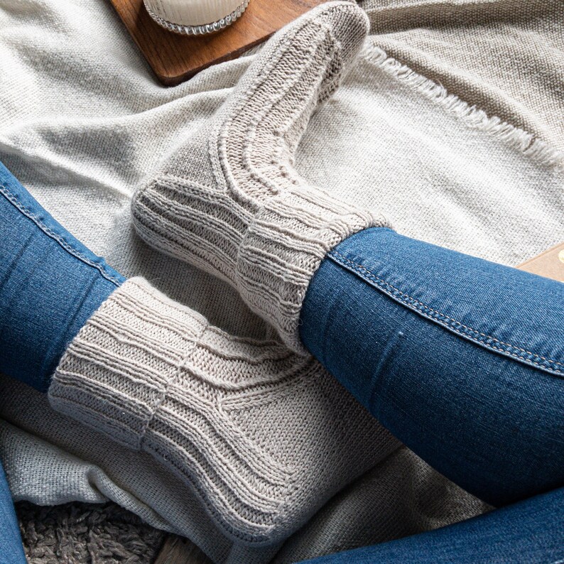 May include: A pair of light gray knitted socks with ribbed cuffs and textured patterns. The socks are worn with blue jeans, suggesting a cozy, casual style. The socks appear to be handmade, emphasizing comfort and warmth.