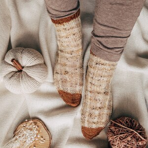 May include: A pair of knitted socks with a brown and cream color pattern. The socks are on a white background with a small white pumpkin and a ball of yarn.