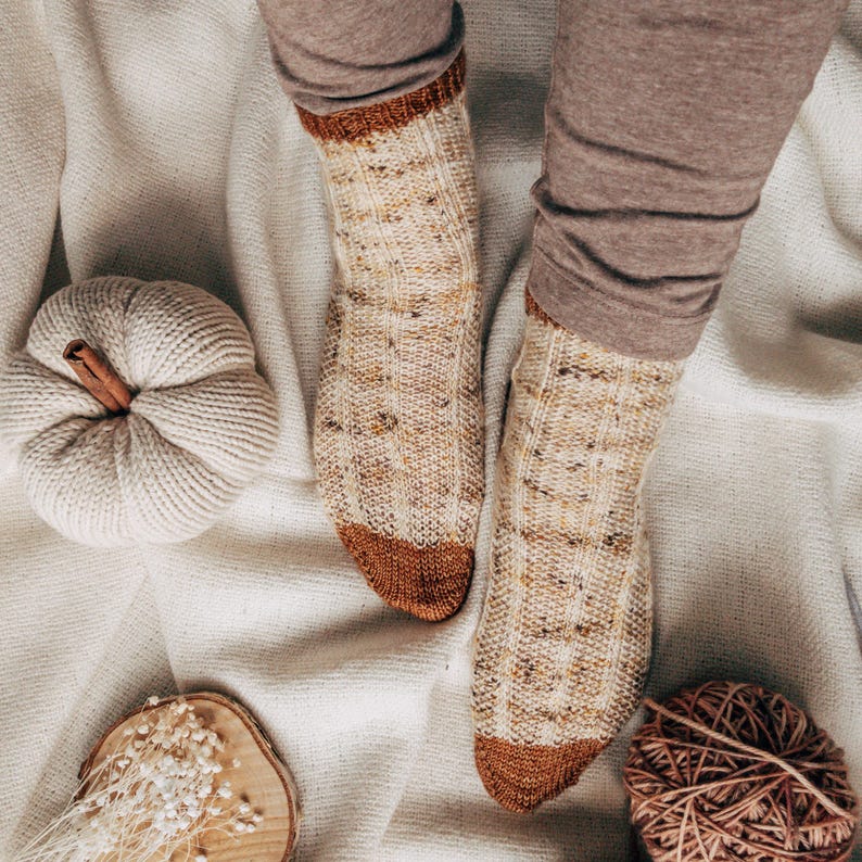 May include: A pair of knitted socks with a brown and cream color pattern. The socks are on a white background with a small white pumpkin and a ball of yarn.