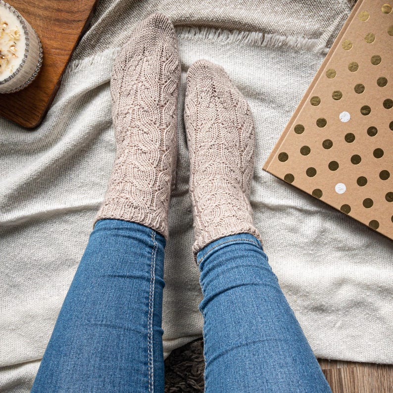 May include: A pair of beige knitted socks with a textured leaf pattern. The socks are worn over blue jeans. A wooden tray with a candle and a gold polka dot gift box are in the background.