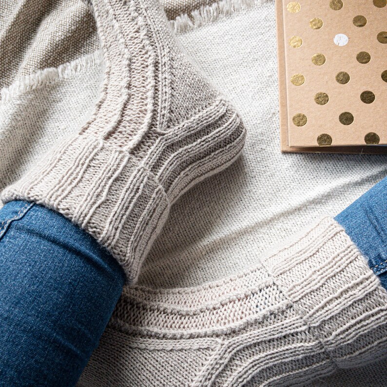 May include: A close-up shot of a person wearing light gray knitted socks with ribbed cuffs and textured patterns. The socks are paired with blue jeans. A gold polka dot card is visible in the background, suggesting a cozy, relaxed setting.