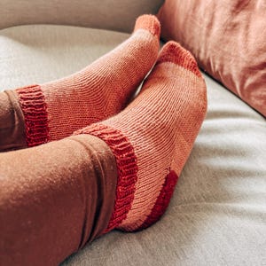 K&ouml;nnte beinhalten: Ein Paar gestrickte Socken in Rosa- und Rott&ouml;nen mit einem gestreiften Muster. Die Socken werden an den F&uuml;&szlig;en einer Person getragen und liegen auf einem hellgrauen Sofa.