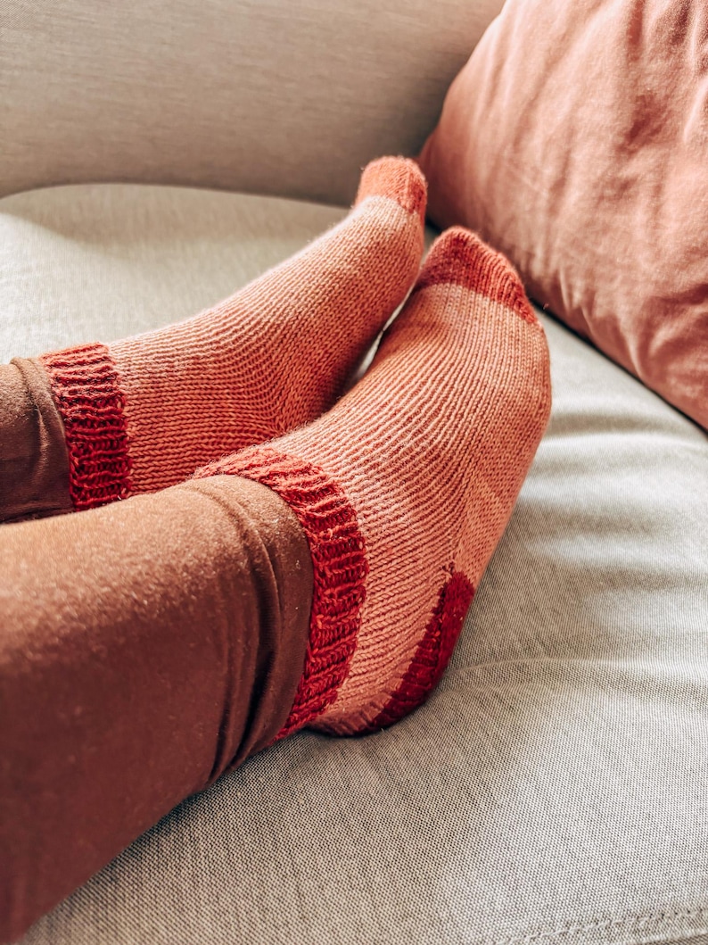 K&ouml;nnte beinhalten: Ein Paar gestrickte Socken in Rosa- und Rott&ouml;nen mit einem gestreiften Muster. Die Socken werden an den F&uuml;&szlig;en einer Person getragen und liegen auf einem hellgrauen Sofa.