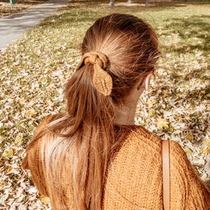 May include: A woman with long, brown hair tied back with a brown knitted hair tie. She is wearing a brown sweater and has a brown leather strap hanging from her shoulder.