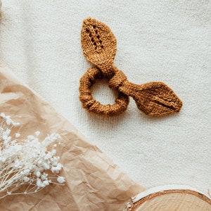 May include: A brown knitted hair scrunchie with two small loops on either side. The scrunchie is lying on a white textured surface.