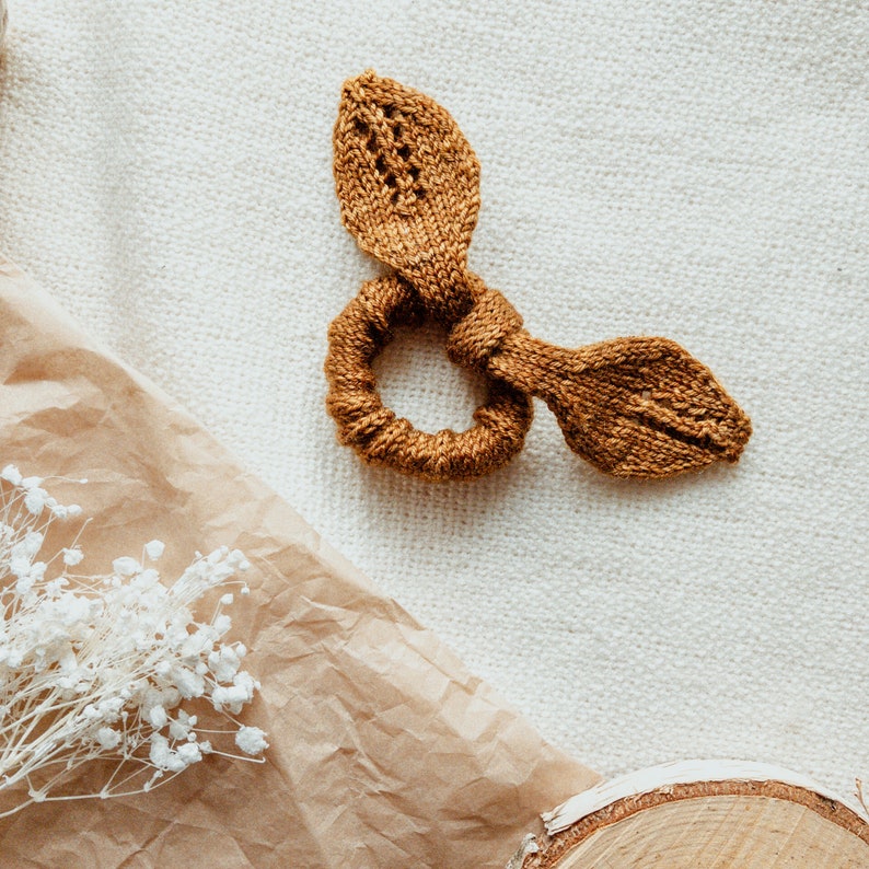 May include: A brown knitted hair scrunchie with two small loops on either side. The scrunchie is lying on a white textured surface.