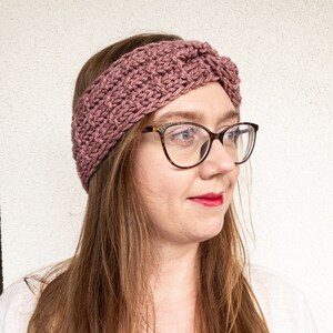 May include: A mauve knitted headband with a twisted design. The headband is worn by a person with long brown hair and glasses.