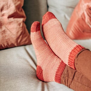 K&ouml;nnte beinhalten: Ein Paar rosa und rote gestreifte Socken mit gerippter Struktur. Die Socken werden von einer Person getragen, die auf einem Sofa sitzt.