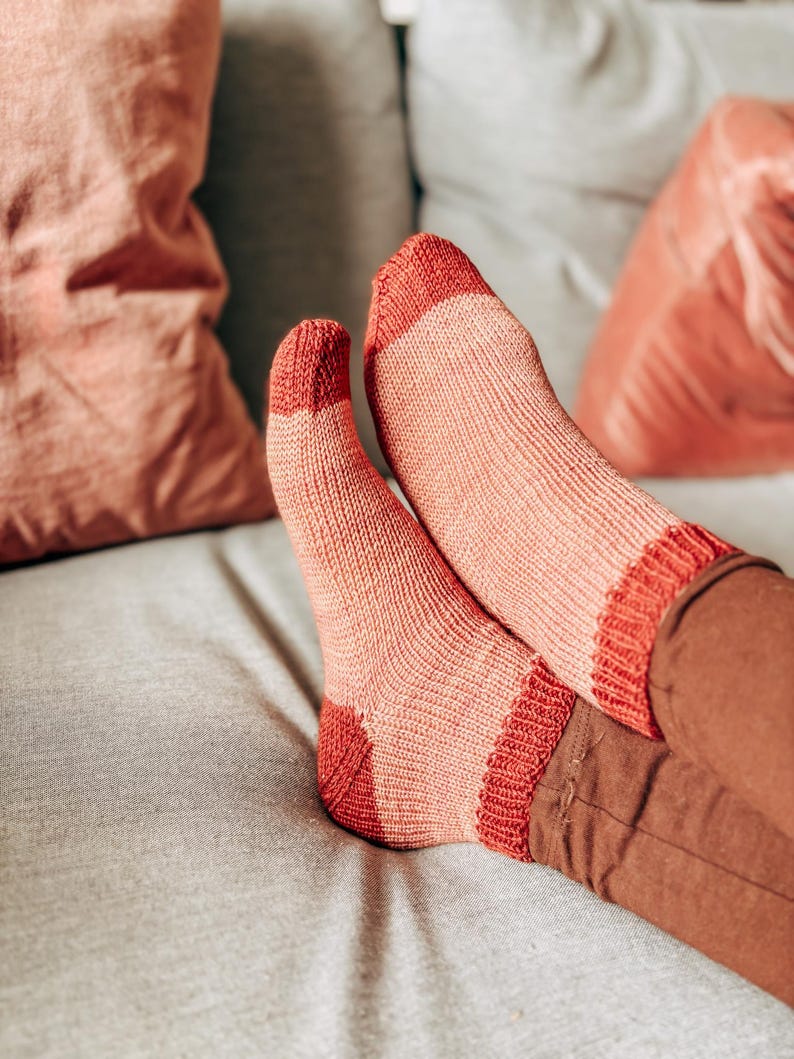 K&ouml;nnte beinhalten: Ein Paar rosa und rote gestreifte Socken mit gerippter Struktur. Die Socken werden von einer Person getragen, die auf einem Sofa sitzt.