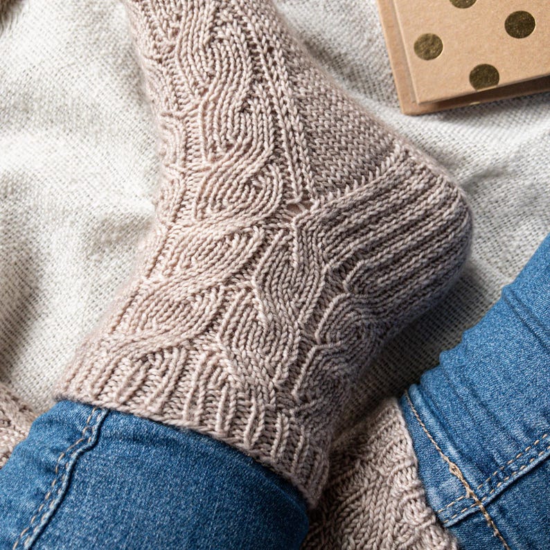 May include: Close-up of a beige cable-knit sock with a textured pattern. The sock is worn with blue denim jeans. A small, gold-dotted brown box is visible in the background. The sock's design features intricate cable patterns.