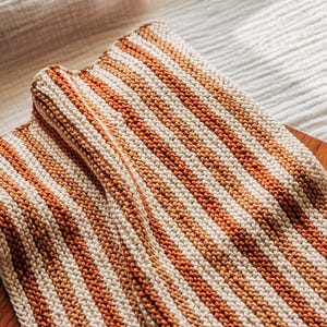May include: A striped knitted blanket in shades of cream and brown. The blanket is made with a simple knit stitch and has a soft, cozy texture.