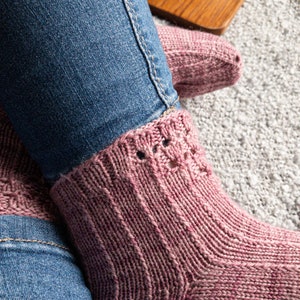 May include: Close-up of a pair of pink knitted socks with a ribbed design and decorative holes at the cuff. The socks are worn with blue jeans. The socks are on a gray carpet.