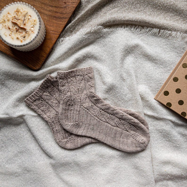 May include: A pair of light brown knitted socks with a cable knit design. The socks are lying on a soft, light gray blanket. A candle in a glass jar and a small gift box with gold polka dots are also in the image.