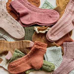 May include: A collection of colorful knitted socks in various patterns and colors, including pink, green, brown, and white. The socks are arranged in a pile, showcasing different textures and styles.